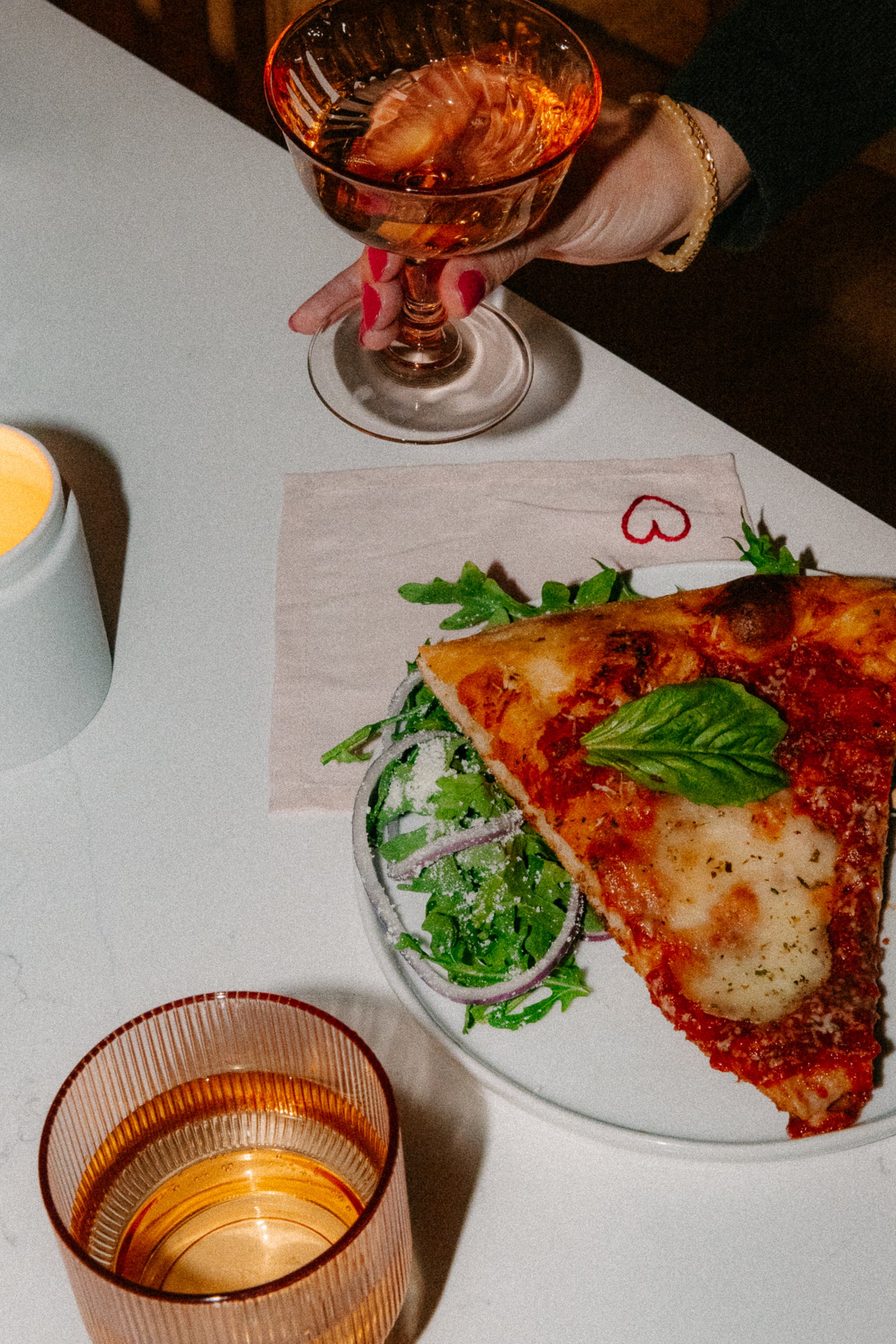 A hand holds a glass of rosé wine above a napkin with a red heart drawing. On the table are a lit candle, another drink, and a plate with two slices of pizza over salad with onions and Parmesan.
