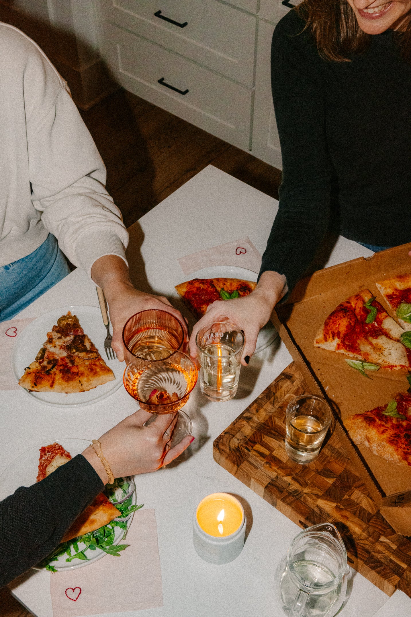 Three people sit around a table sharing pizza, toasting with drinks in glasses. A lit candle and napkins with hearts are on the table, creating a warm, casual atmosphere.