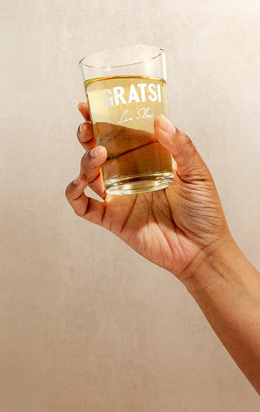 A hand holds a clear glass filled with a light amber liquid. The glass has the word “GRATSI” and “Live Slow” printed on it in white letters against a neutral beige background.