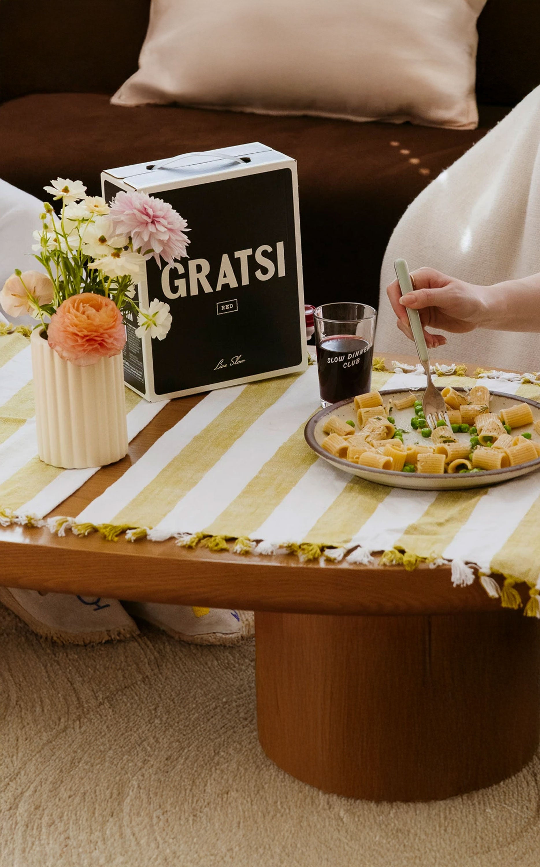 A plate of pasta and a glass are on a wooden table with a striped yellow and white tablecloth, next to a vase of colorful flowers and a Gratsi wine box. A person’s hand holds a fork over the plate.