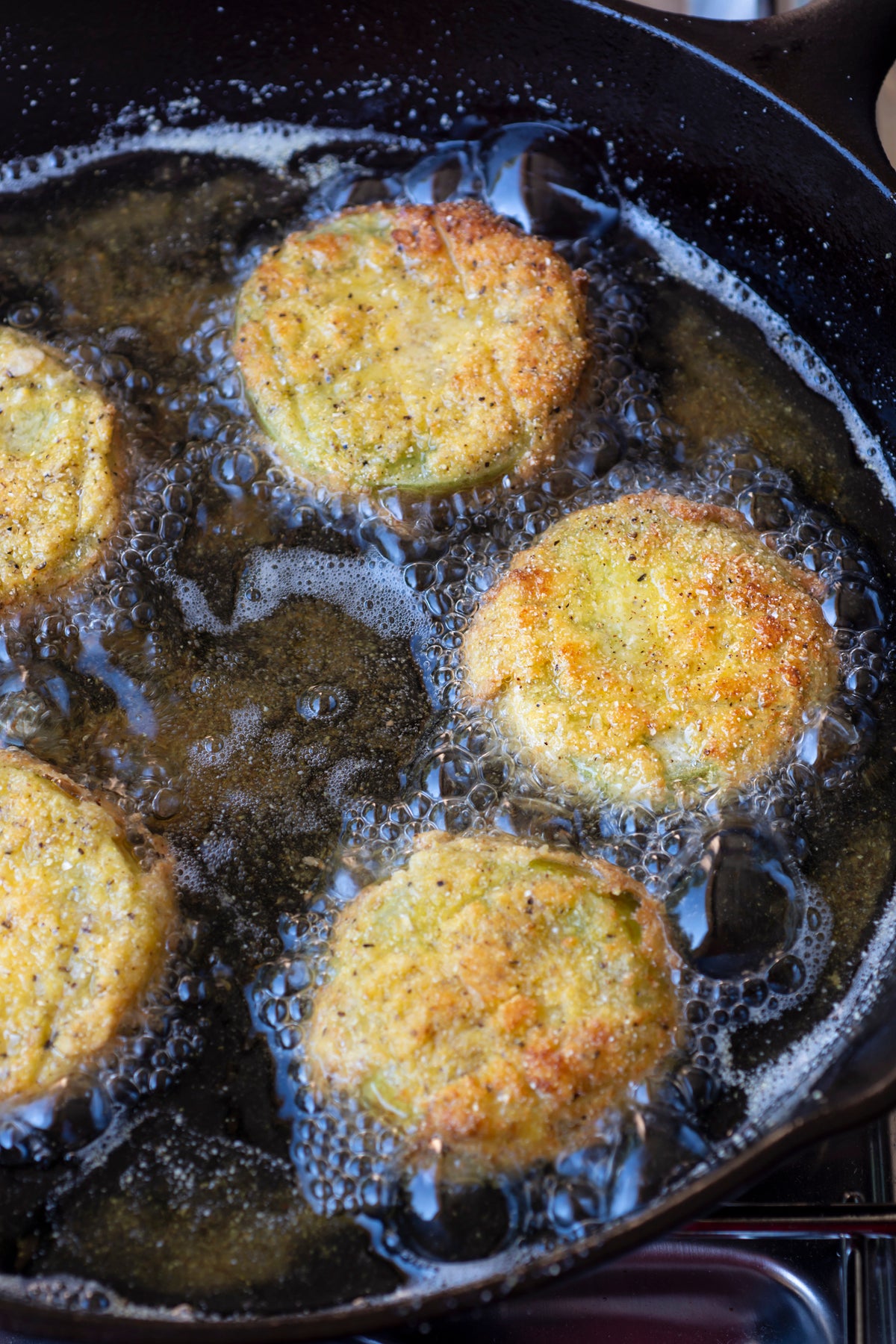 Golden slices of breaded green tomatoes frying in hot oil in a black skillet, with bubbles forming around the edges.