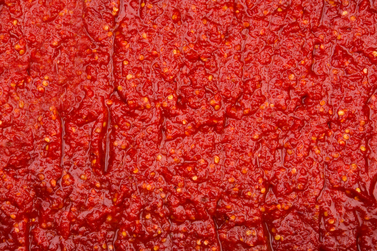 Close-up view of a textured red chili paste with visible chili seeds and pieces, creating a vibrant, spicy appearance.