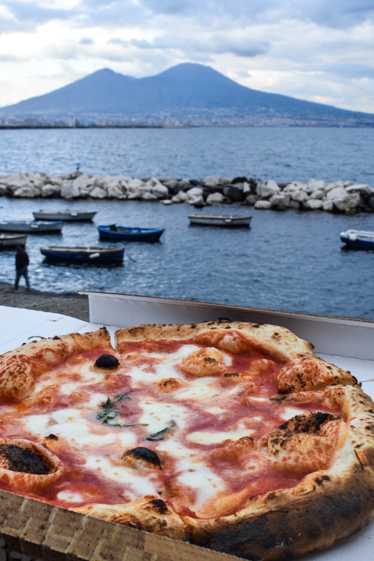 A Neapolitan pizza in an open box sits on a ledge by the sea, with boats on the water and Mount Vesuvius in the background under a cloudy sky.