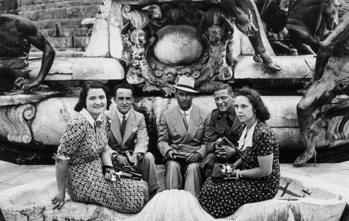 Five people in formal 1930s clothing sit in front of an ornate stone fountain decorated with statues. Three men in suits and two women in patterned dresses pose for the photo, looking toward the camera.