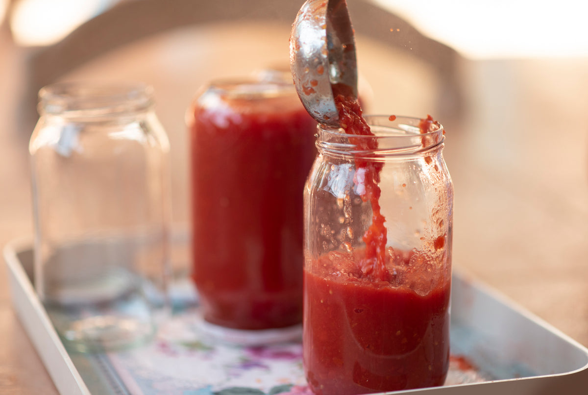 A metal ladle pours red tomato sauce into a glass jar, with two more jars—one filled and one empty—on a tray in the background.