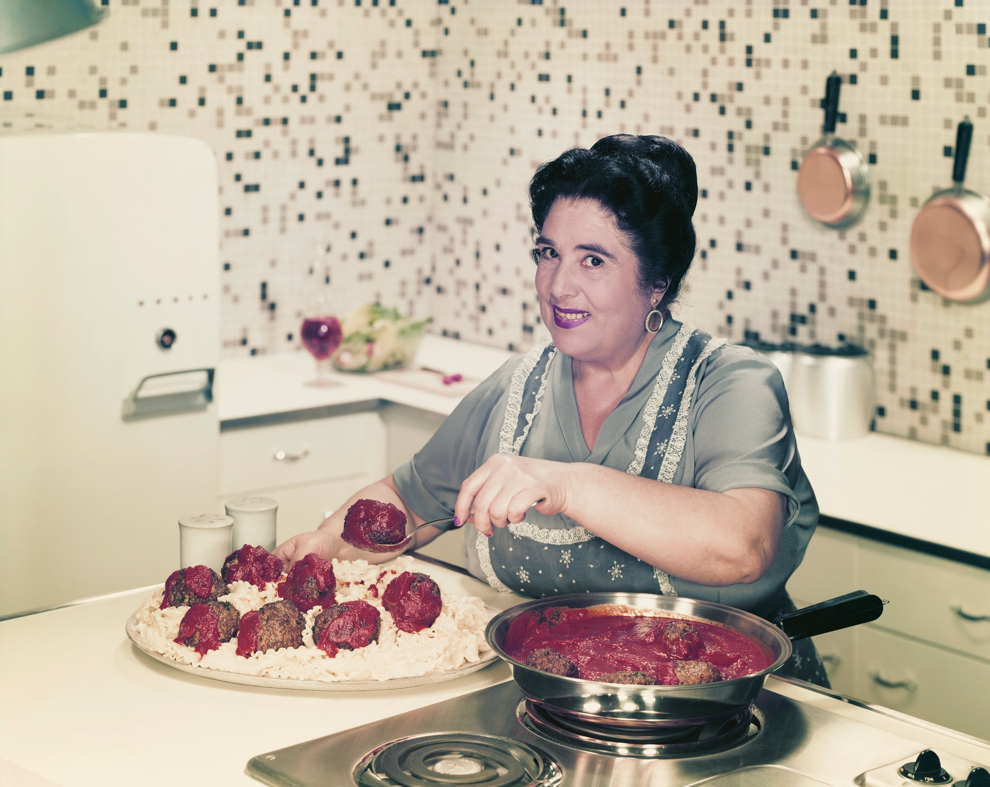 A woman in a vintage kitchen smiles while serving meatballs in red sauce onto a plate of pasta. Pots hang on a tiled wall, and a skillet sits on the stovetop.
