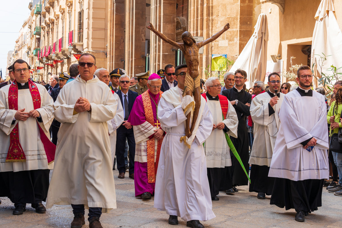 A group of clergy and people participate in a religious procession, with one person carrying a crucifix and others dressed in robes, walking through a street lined with buildings.
