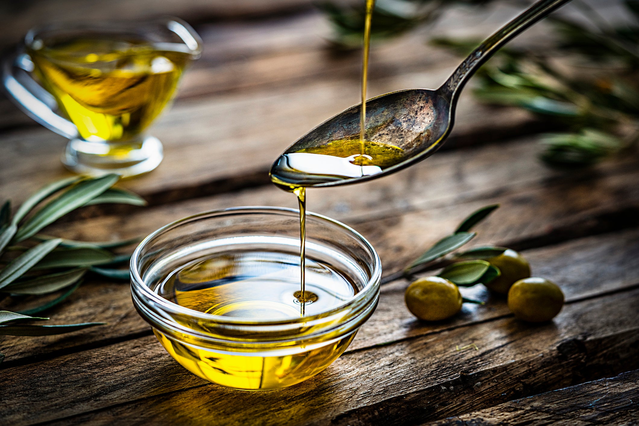 A spoon drizzles golden olive oil into a small glass bowl. Olive branches, whole olives, and a glass jug with more oil sit on a rustic wooden table.