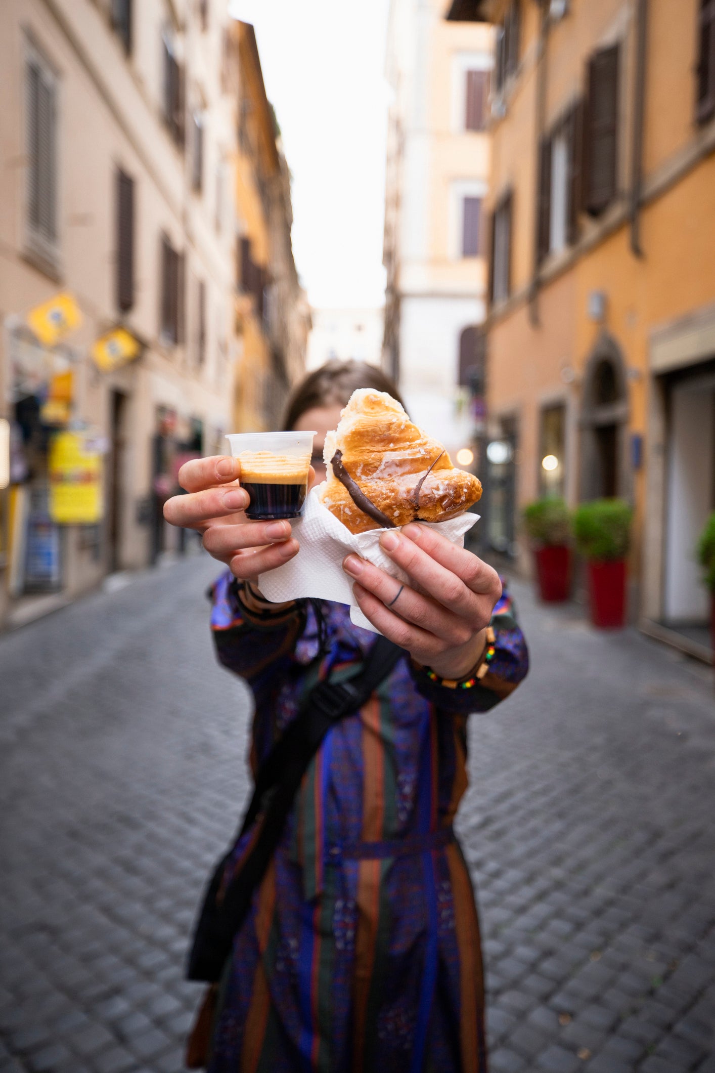 A person stands on a cobblestone street, holding a croissant and a small cup of espresso towards the camera, obscuring their face. The background shows colorful buildings and potted plants.