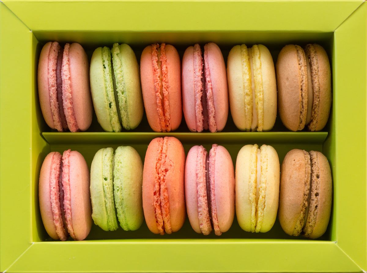 A box of twelve colorful macarons arranged in two rows, with flavors in shades of pink, green, orange, yellow, and brown, inside a light green box.