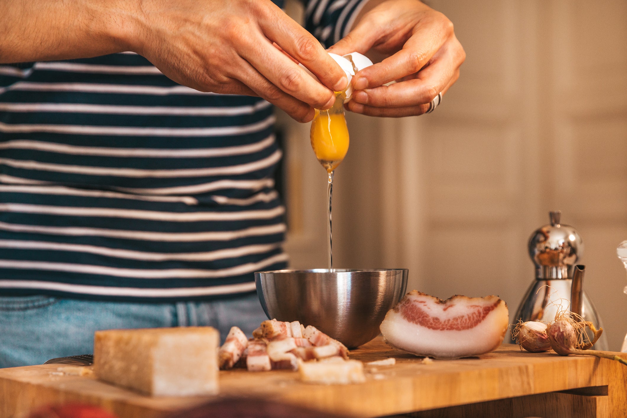 A person in a striped shirt cracks an egg into a metal bowl on a wooden cutting board, surrounded by chopped meat, cheese, garlic, and a pepper mill.