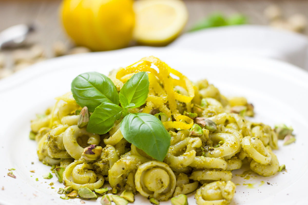 Pasta with green pesto sauce, garnished with fresh basil leaves, lemon zest, and chopped pistachios, served on a white plate. A sliced lemon and scattered pistachios are in the blurred background.
