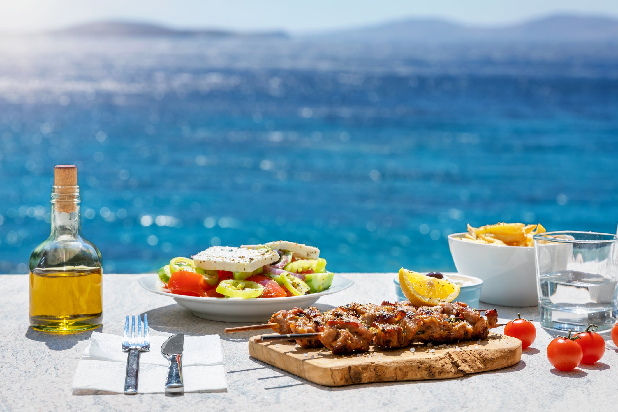 A Greek meal with salad, skewered meat, fries, olive oil, and water is set on a table overlooking the blue sea and distant hills under a sunny sky.
