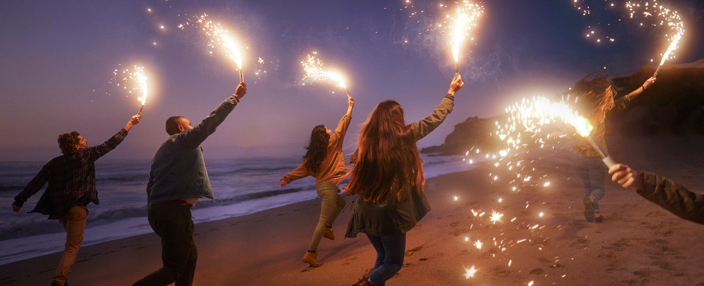 Five people run along a beach at dusk, holding sparklers high in the air, with the ocean and cliffs visible in the background. The sky is dusky purple, creating a festive and joyful atmosphere.