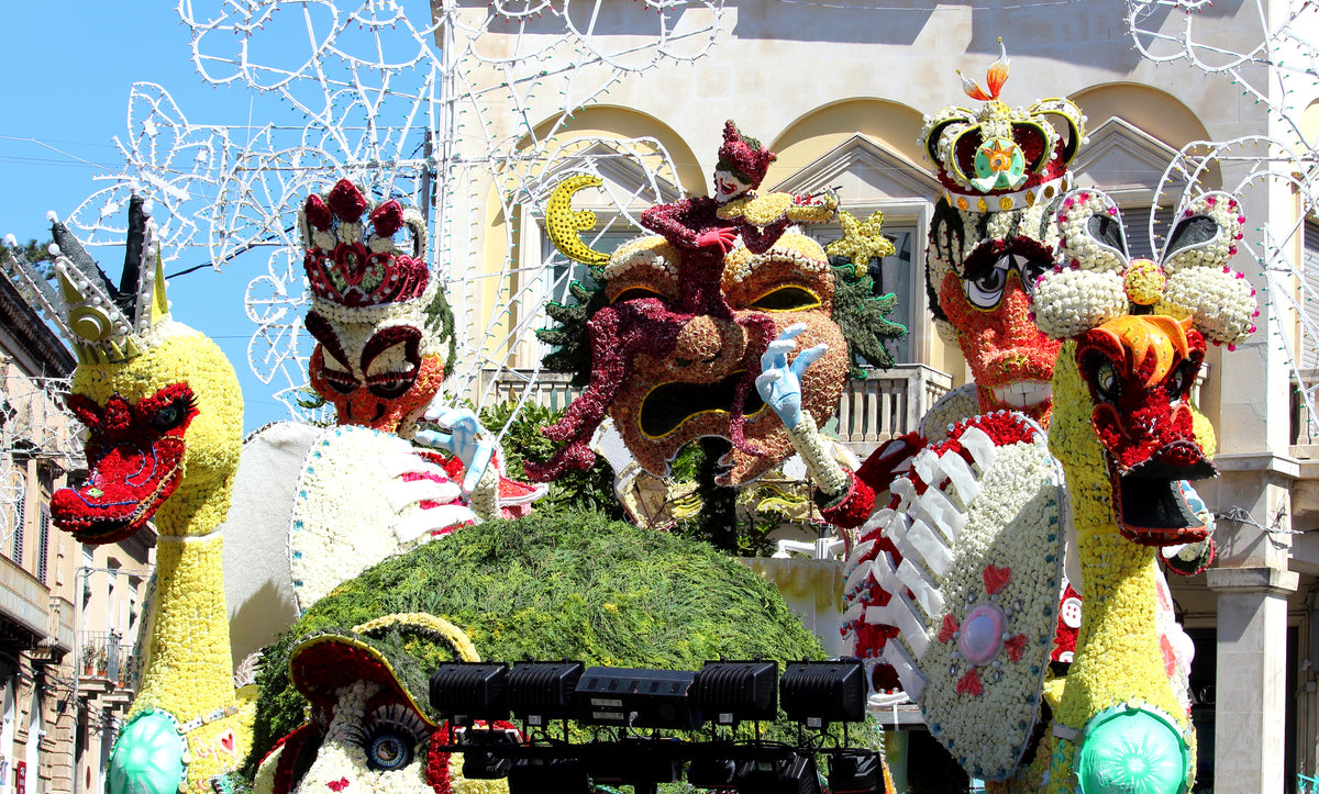 Colorful parade floats decorated with elaborate masks, crowns, dragons, and flowers stand against a sunny building backdrop. White decorative lights hang above, and bright yellow birds are in the foreground.