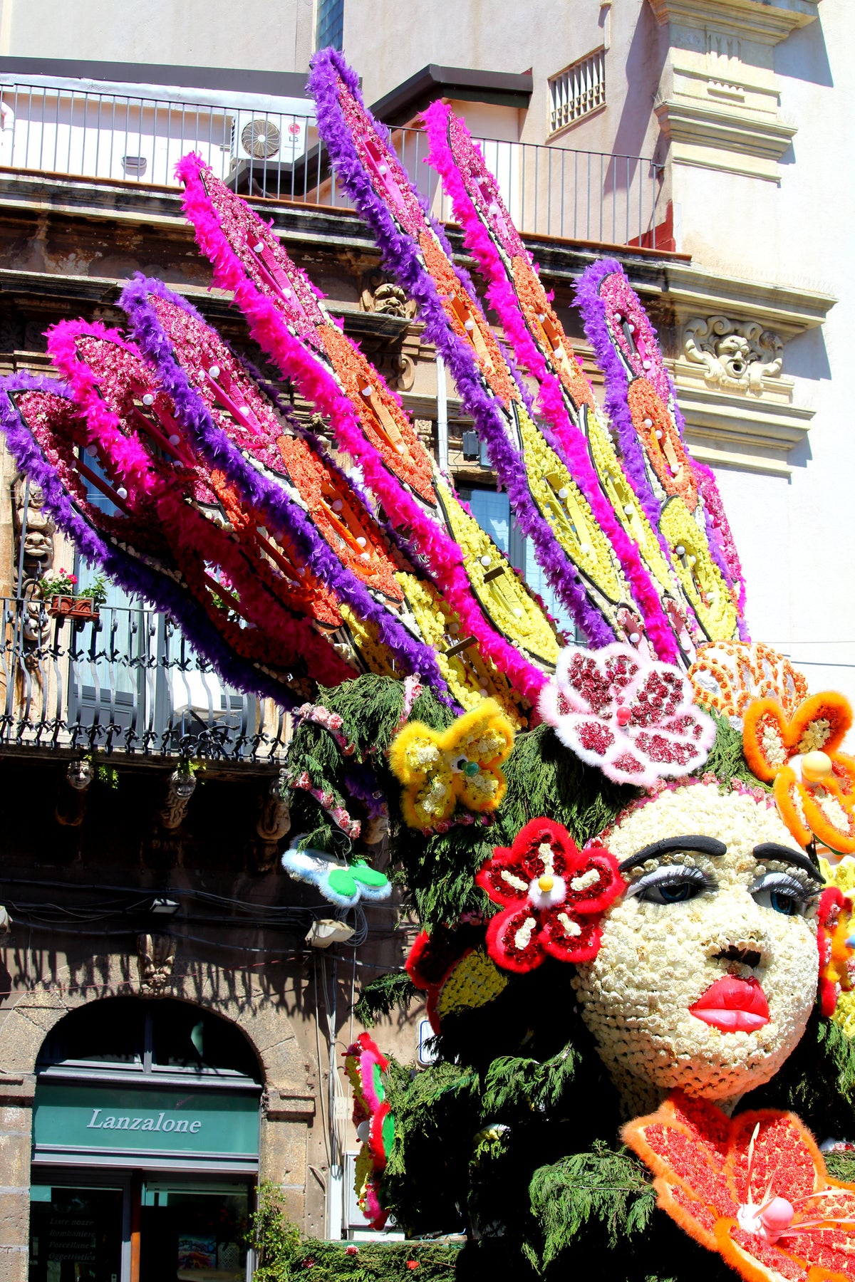 A large parade float features a womans face made of flowers and greenery, with vibrant, colorful floral headpieces and bright red lips, set against a sunny cityscape.
