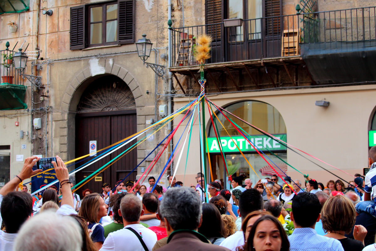 A crowd gathers as people dance around a maypole with colorful ribbons in a lively outdoor celebration, set against old buildings and a pharmacy storefront.