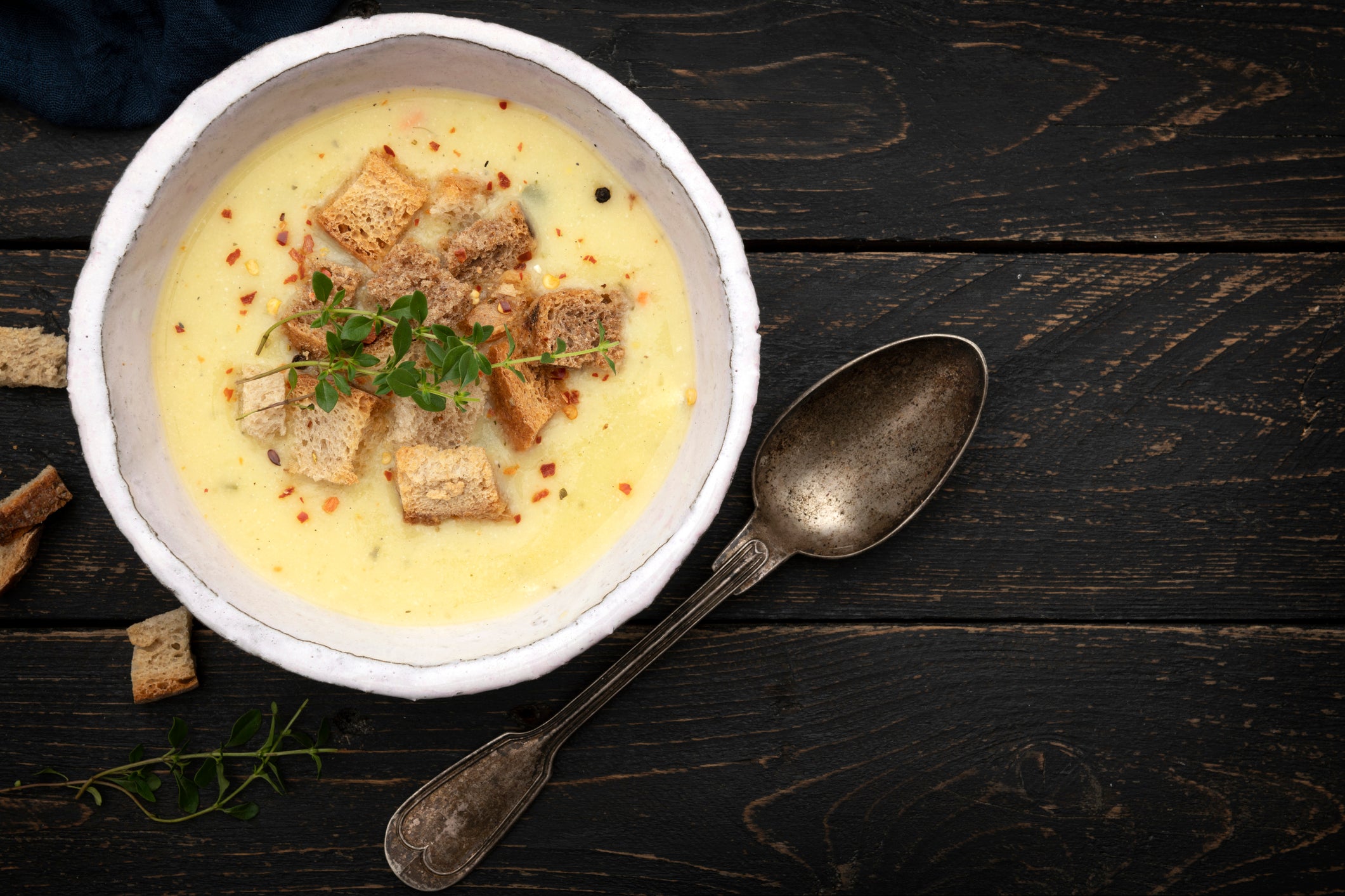 A bowl of creamy soup topped with croutons and herbs sits on a dark wooden table. An antique spoon rests beside the bowl, and some croutons and herbs are scattered nearby.