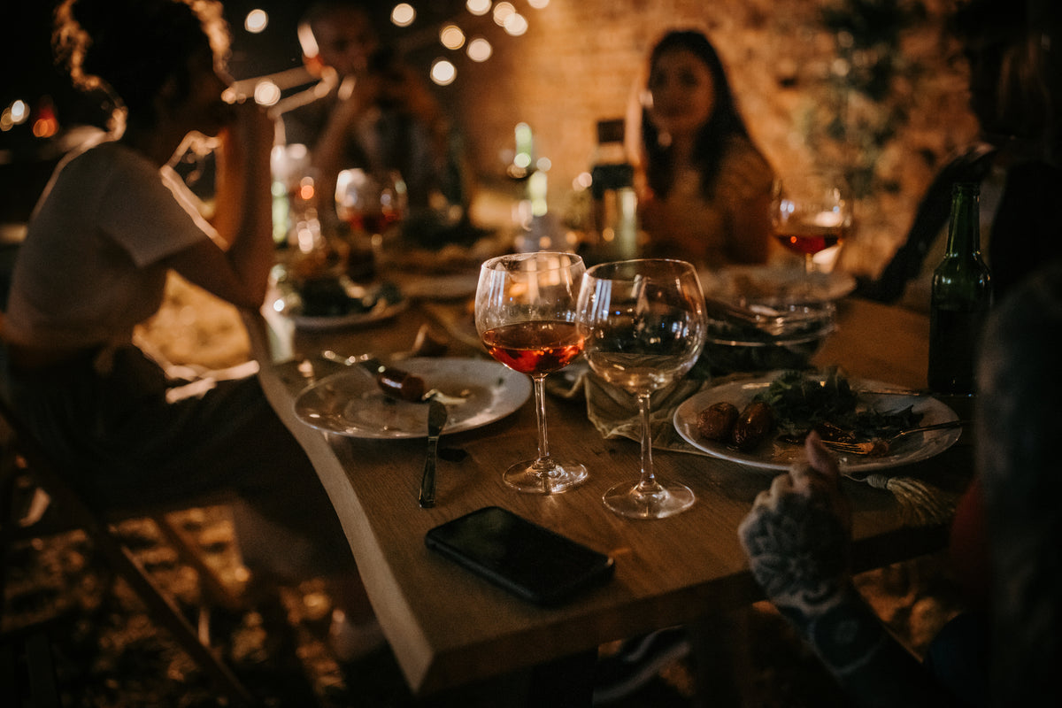 A group of people sit around a table outdoors at night, enjoying food and drinks. The scene is dimly lit with string lights, creating a warm, festive atmosphere.