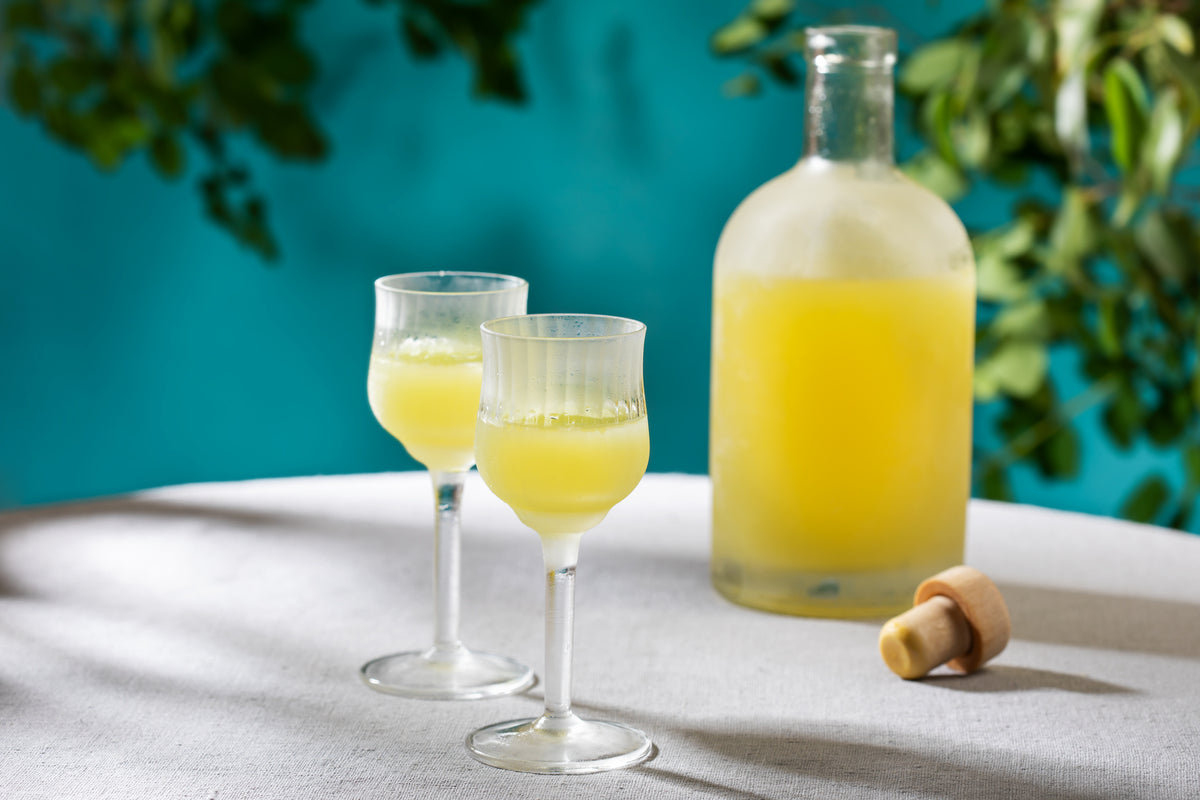 A frosted glass bottle of yellow liqueur with a cork beside it and two filled stemmed glasses sit on a white tablecloth, with a blue background and green leaves in soft focus.