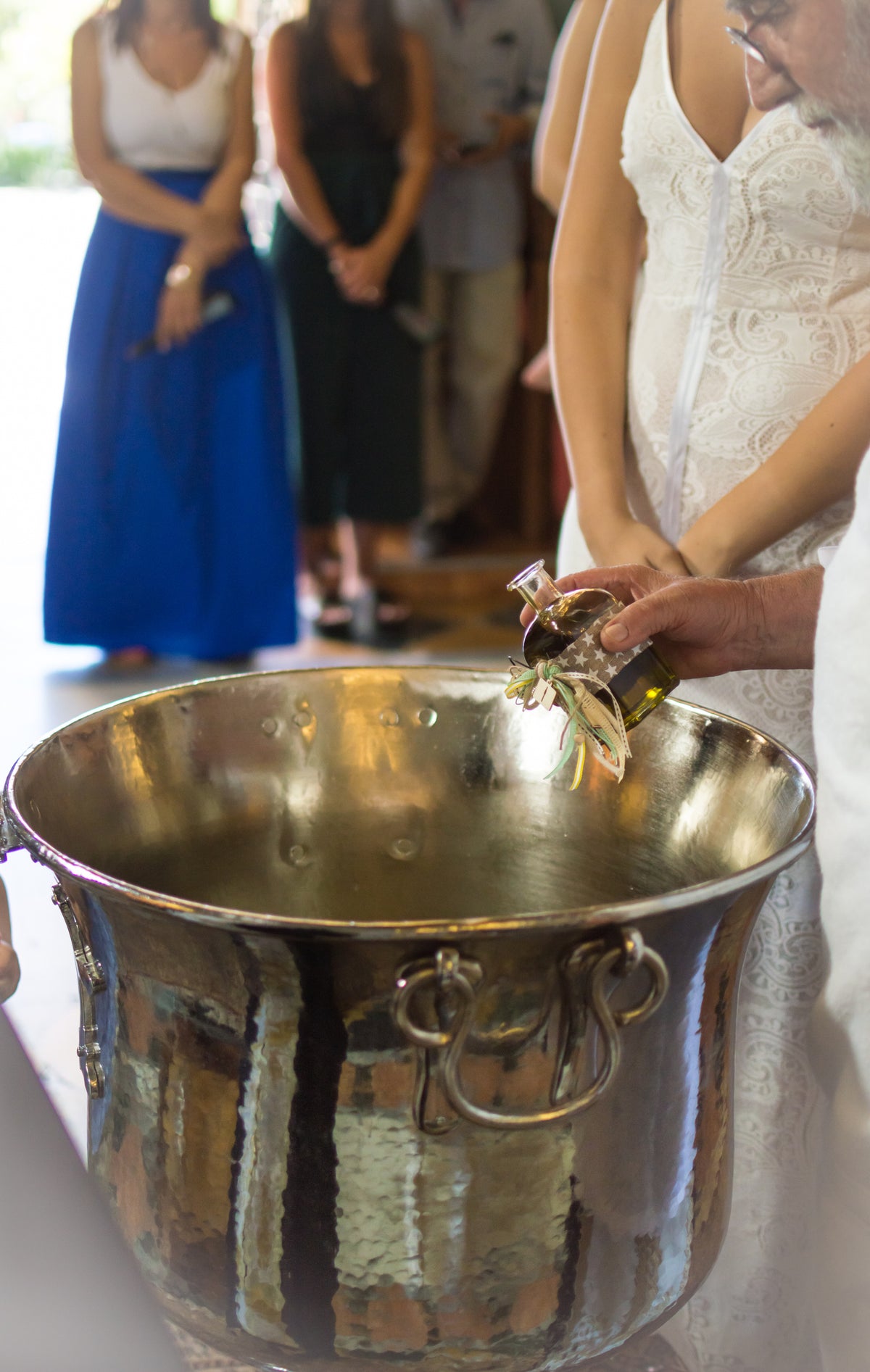A person holds a small ornate cross over a large silver basin, with people dressed formally standing around, suggesting a religious ceremony.