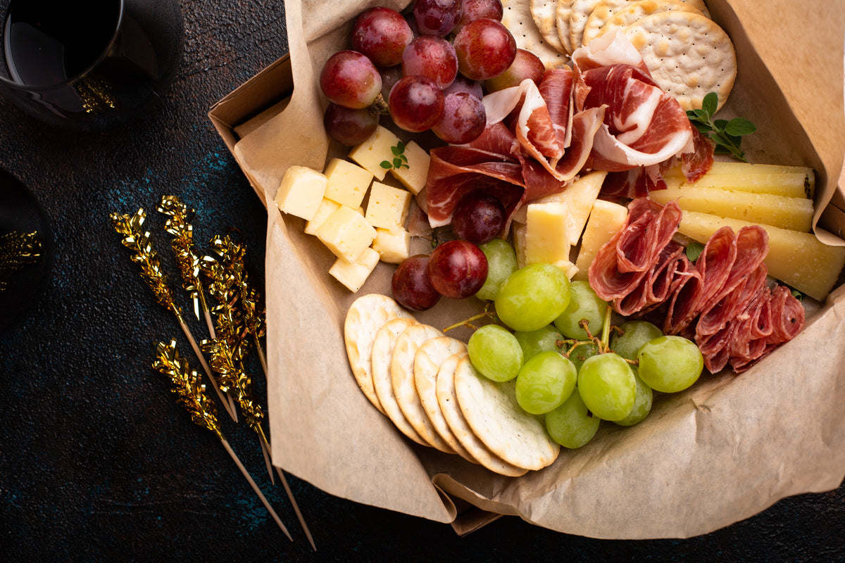 A charcuterie box with red and green grapes, sliced cured meats, various cheeses, round crackers, and bread, arranged neatly in brown paper. Two drinks and gold-decorated stems are beside the box on a dark surface.