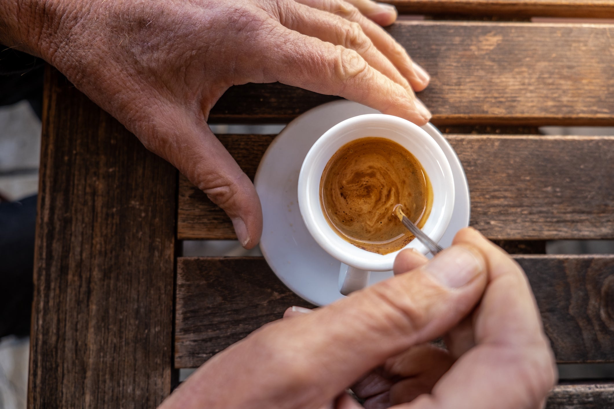 A person stirs a cup of espresso with a spoon on a wooden table, with another hand resting nearby.