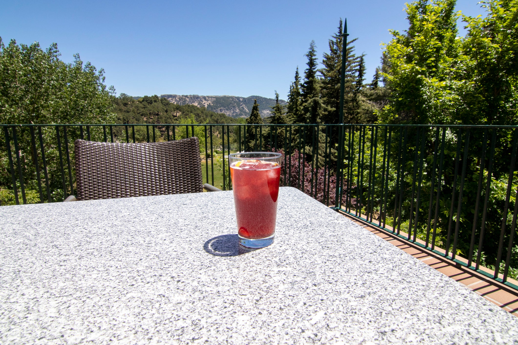 A glass of red fruit drink with ice sits on a stone table outdoors, with a wicker chair and scenic trees and mountains in the background under a clear blue sky.