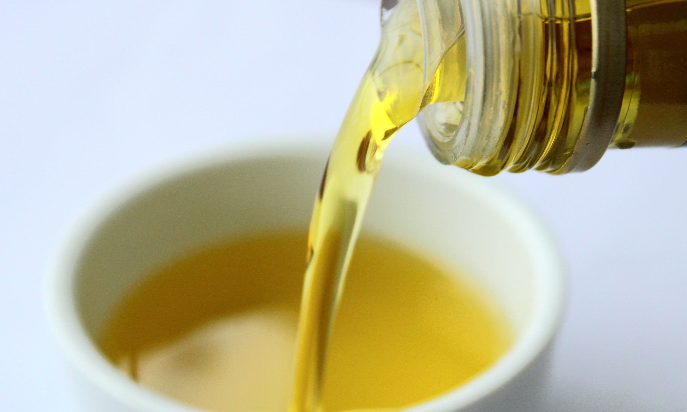 Golden-yellow oil being poured from a bottle into a white bowl, with the oil midstream and close-up detail on the bottles neck and the liquid surface in the bowl.