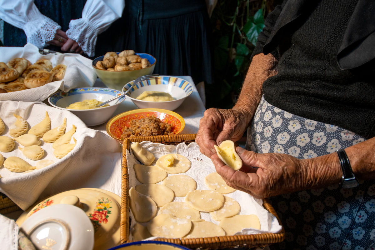 An elderly woman fills and folds dough by hand at a table with bowls of ingredients and uncooked pastries, preparing traditional food.