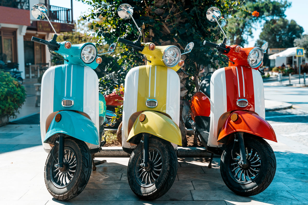 Three colorful scooters—blue, yellow, and orange—are parked side by side on a sunny sidewalk, with greenery and buildings visible in the background.