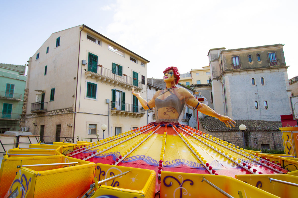 A colorful carnival ride with a large figure of a woman at its center stands in front of old, multi-story buildings with green shutters and balconies under a partly cloudy sky.