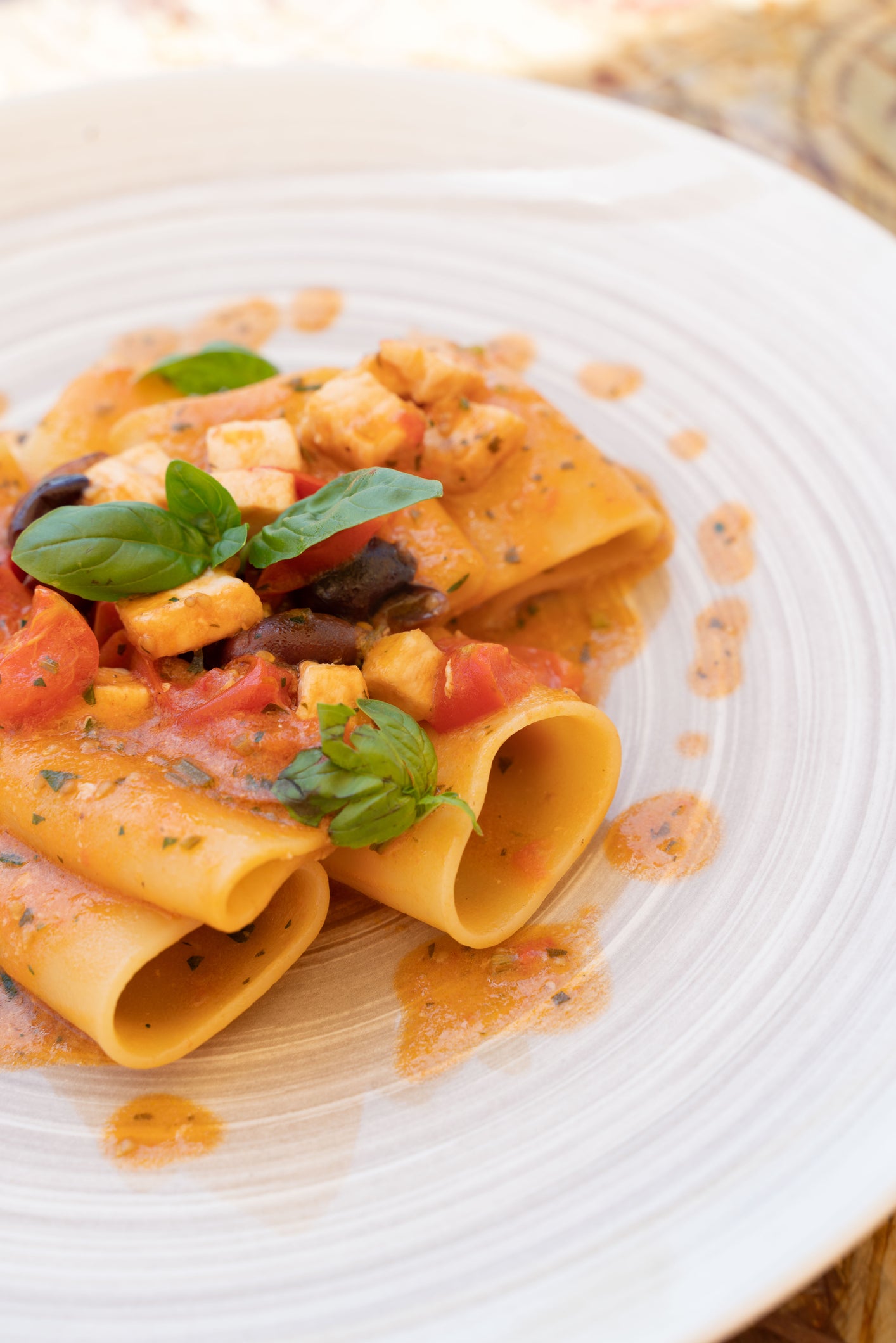A close-up of pasta tubes topped with tomato sauce, diced cheese, olives, and fresh basil, served on a white plate.