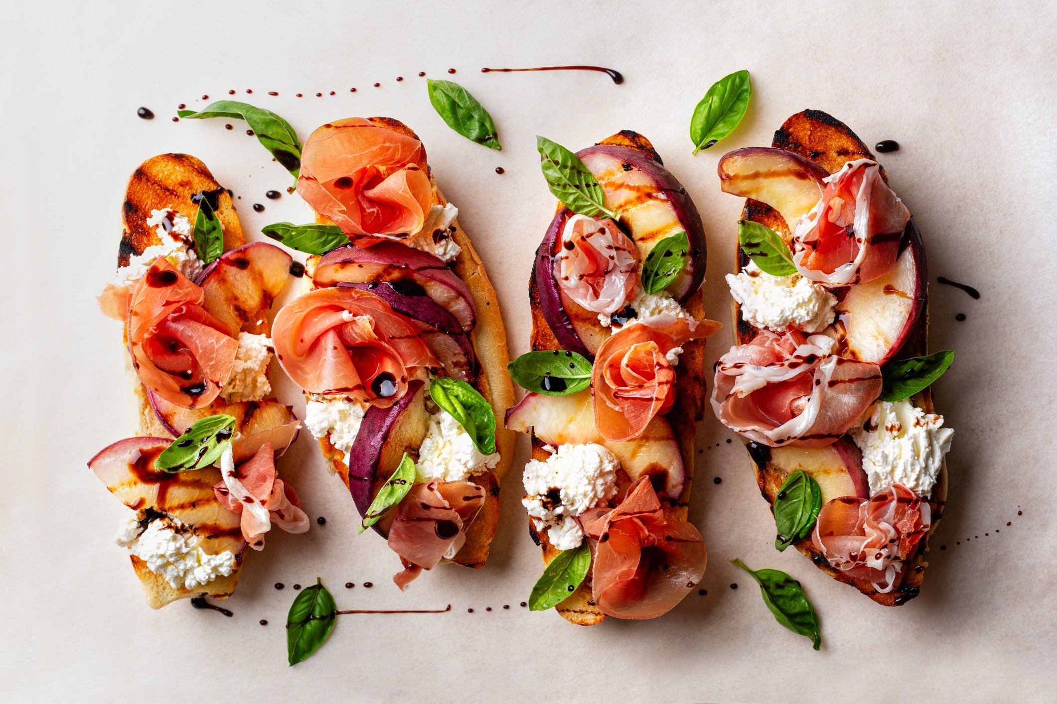 Three pieces of toasted bread topped with ricotta, prosciutto, peach slices, basil leaves, and a drizzle of balsamic glaze, arranged on parchment paper.