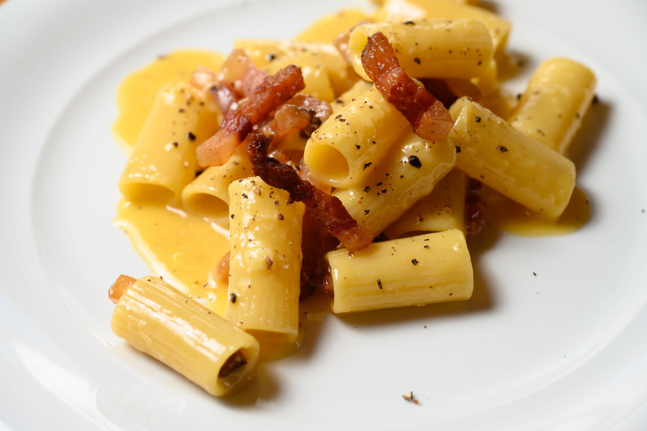 Pasta rigatoni with creamy yellow sauce, crispy bacon pieces, and cracked black pepper, served on a white plate.