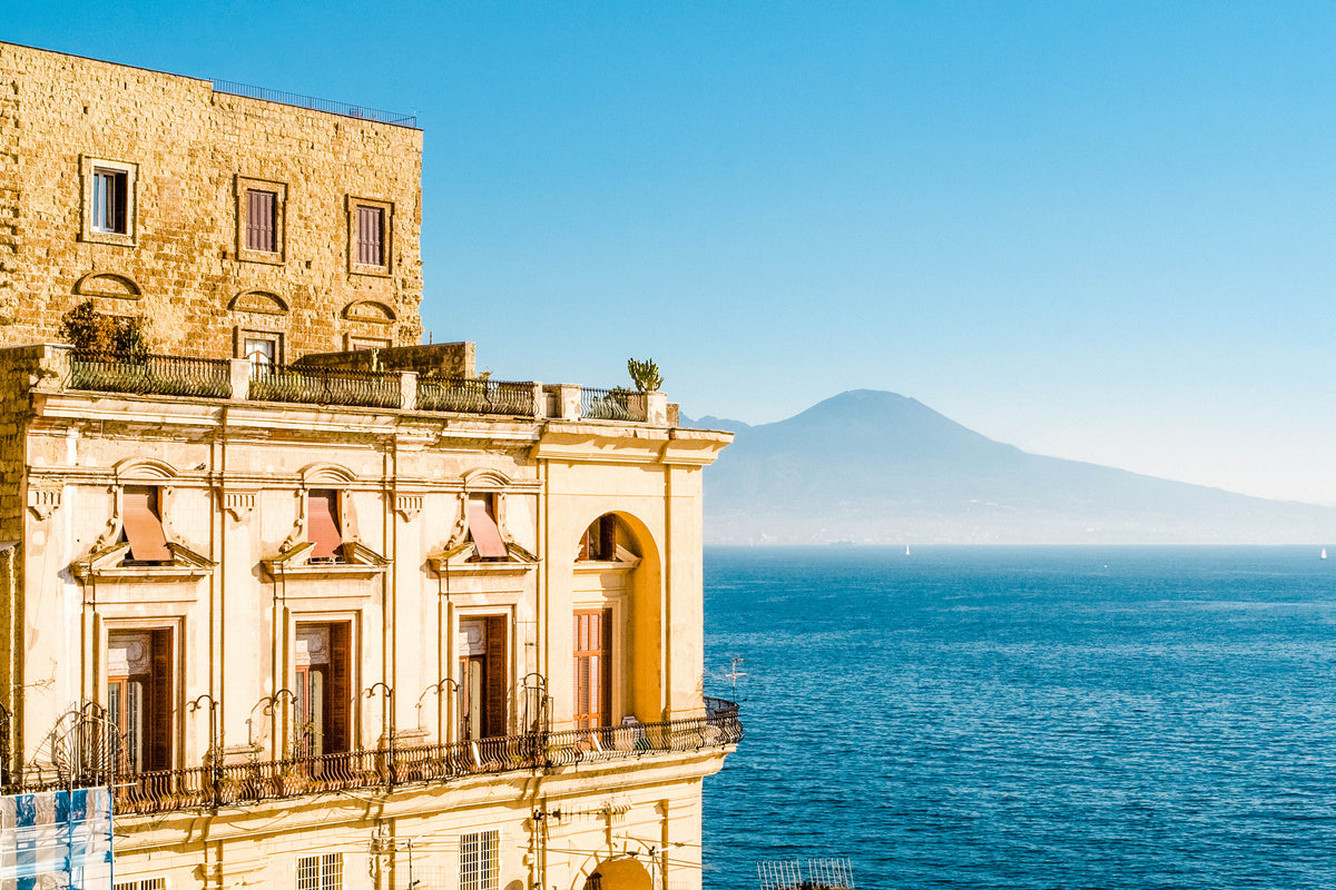 A yellow historic building overlooks a blue sea, with distant sailboats and a large mountain visible under a clear blue sky in the background.