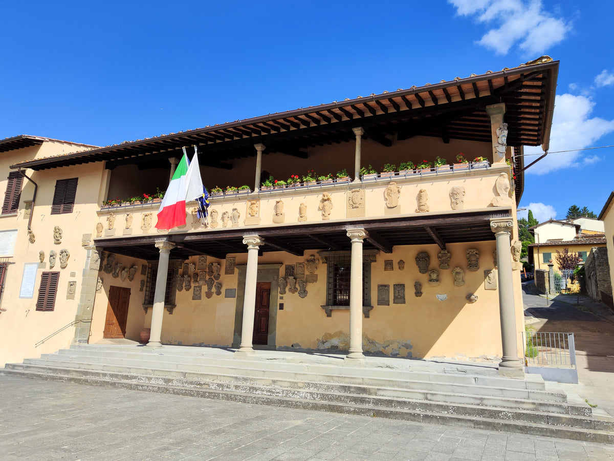 A historic building with columns, wall plaques, and flower boxes. The Italian and European Union flags hang from the balcony. The sky is blue with scattered clouds.