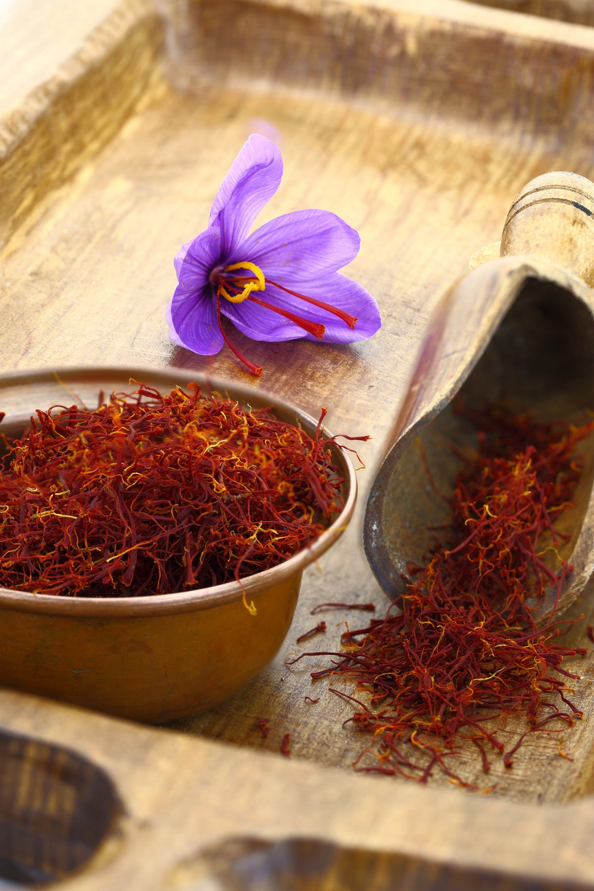 A purple saffron flower, a brass bowl filled with saffron threads, and a wooden scoop with spilled saffron threads on a rustic wooden surface.