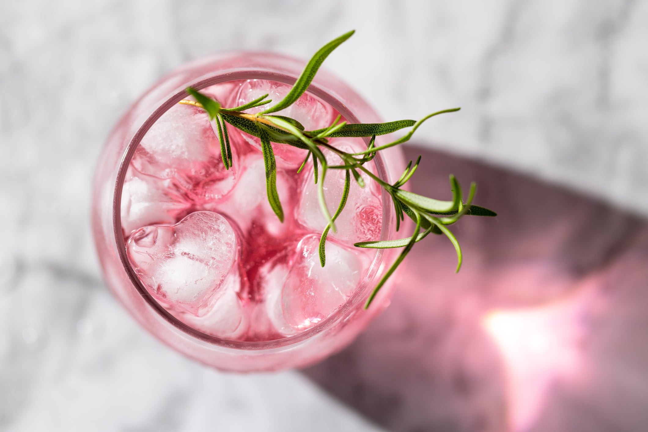 A pink drink with ice cubes and a sprig of fresh rosemary in a glass, viewed from above, sits on a light marble surface with a shadow and light reflection.