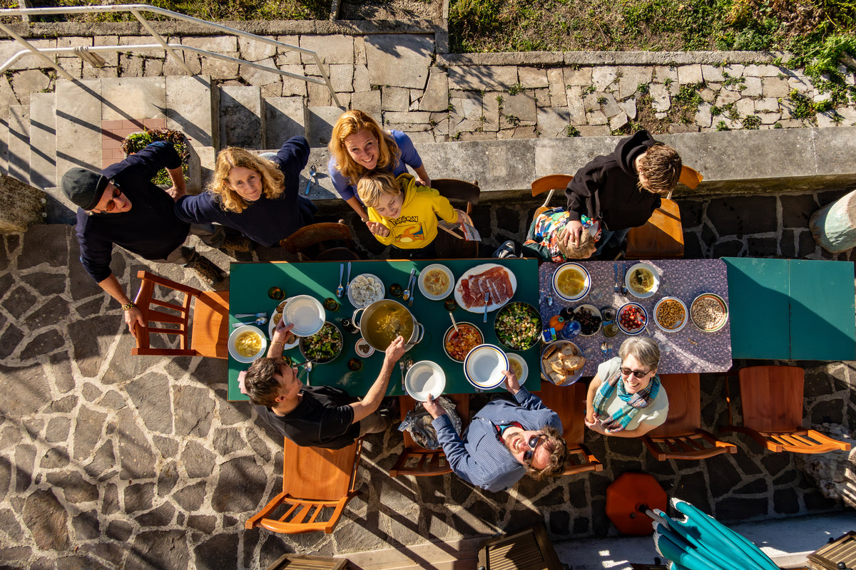 A group of people gathered around a long outdoor table set with various dishes, smiling and looking up toward the camera on a sunny day.