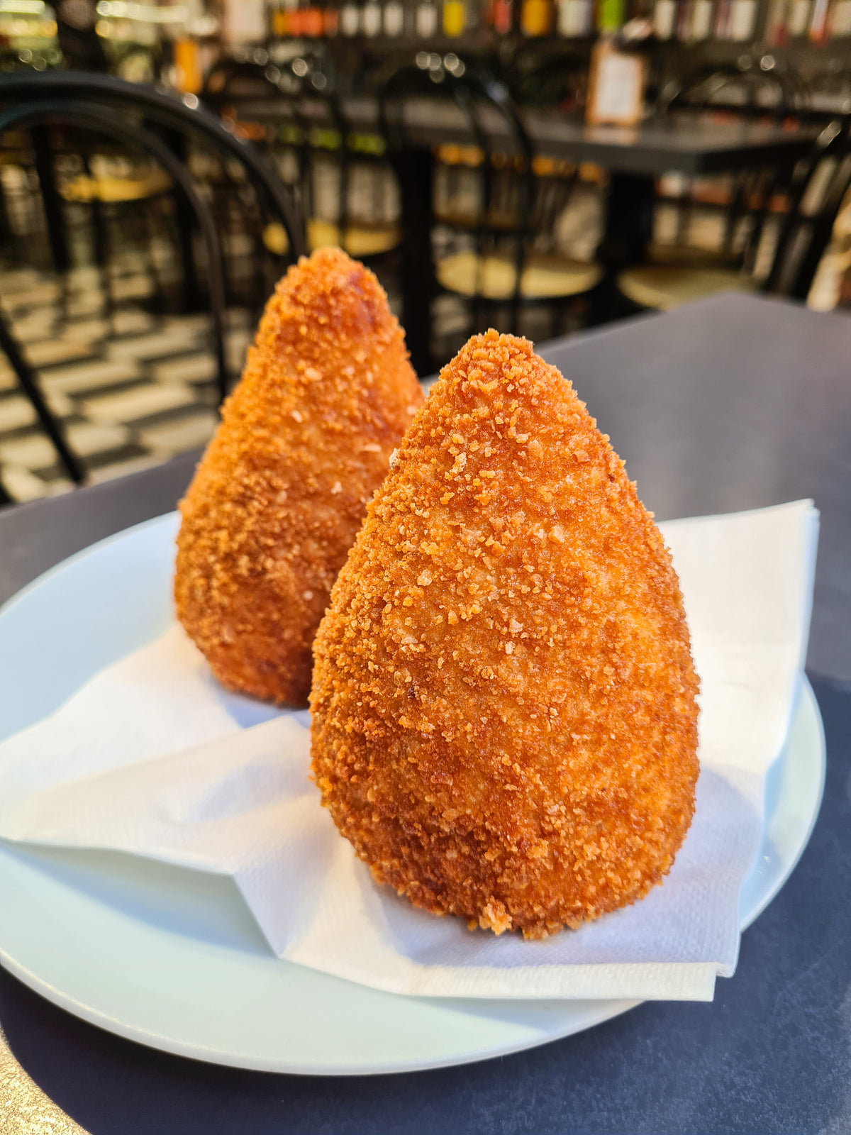 Two golden, cone-shaped fried snacks sit on a white plate lined with napkins, with a blurred café setting in the background.