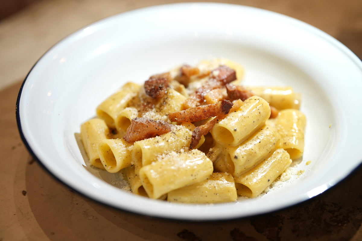 A white plate with rigatoni pasta in a creamy yellow sauce, garnished with crispy bacon pieces and sprinkled with grated cheese and black pepper.