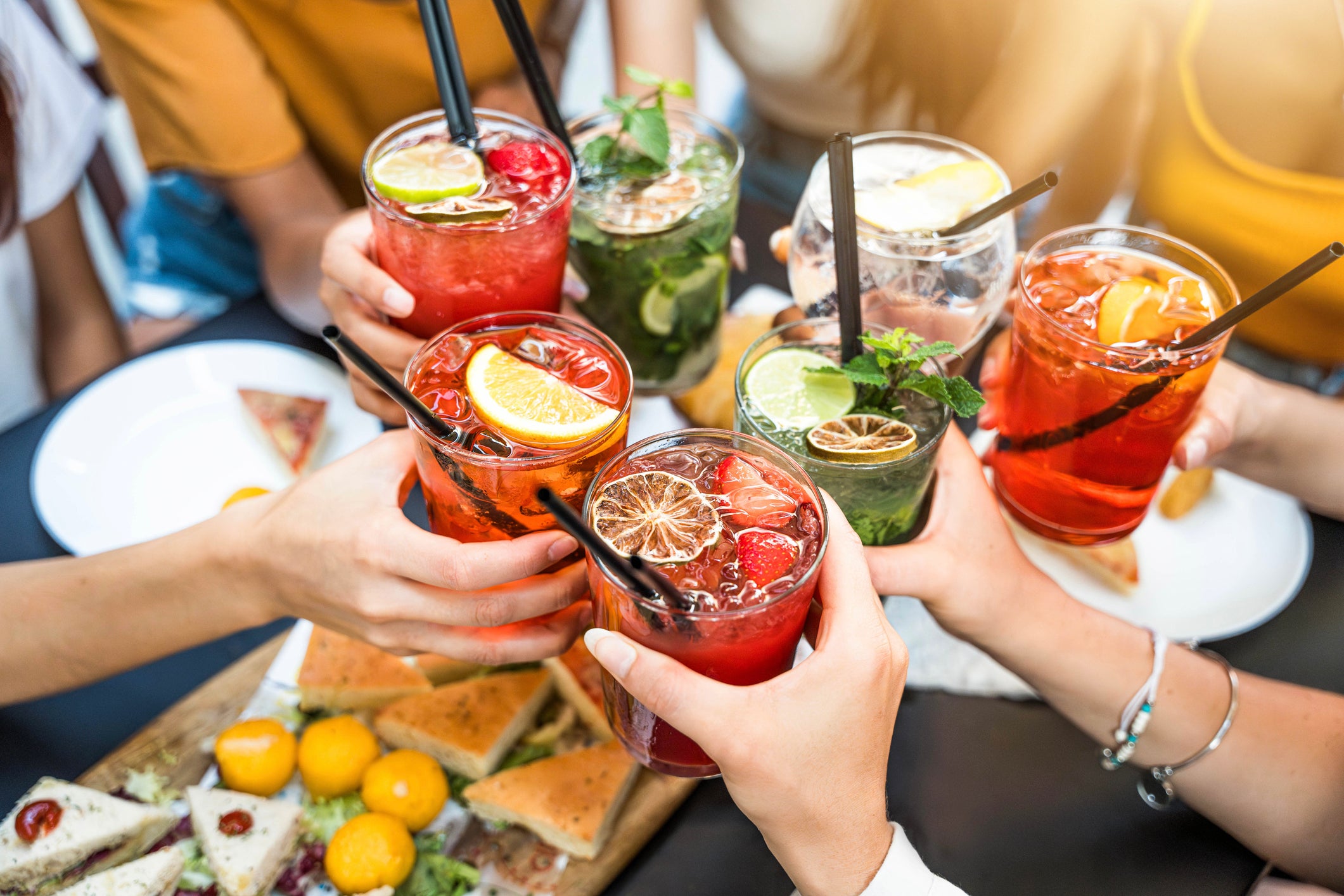 A group of people toast with colorful cocktails garnished with citrus and mint, above a table with assorted appetizers and snacks.