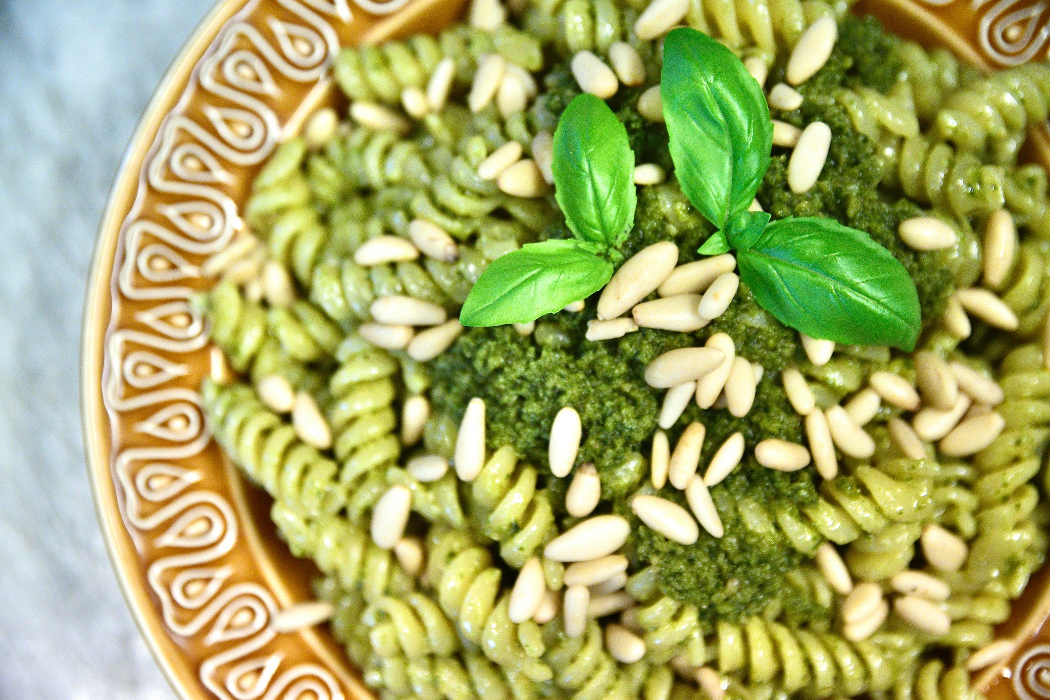 A close-up of rotini pasta with green pesto sauce, topped with pine nuts and fresh basil leaves, served in a decorative bowl.