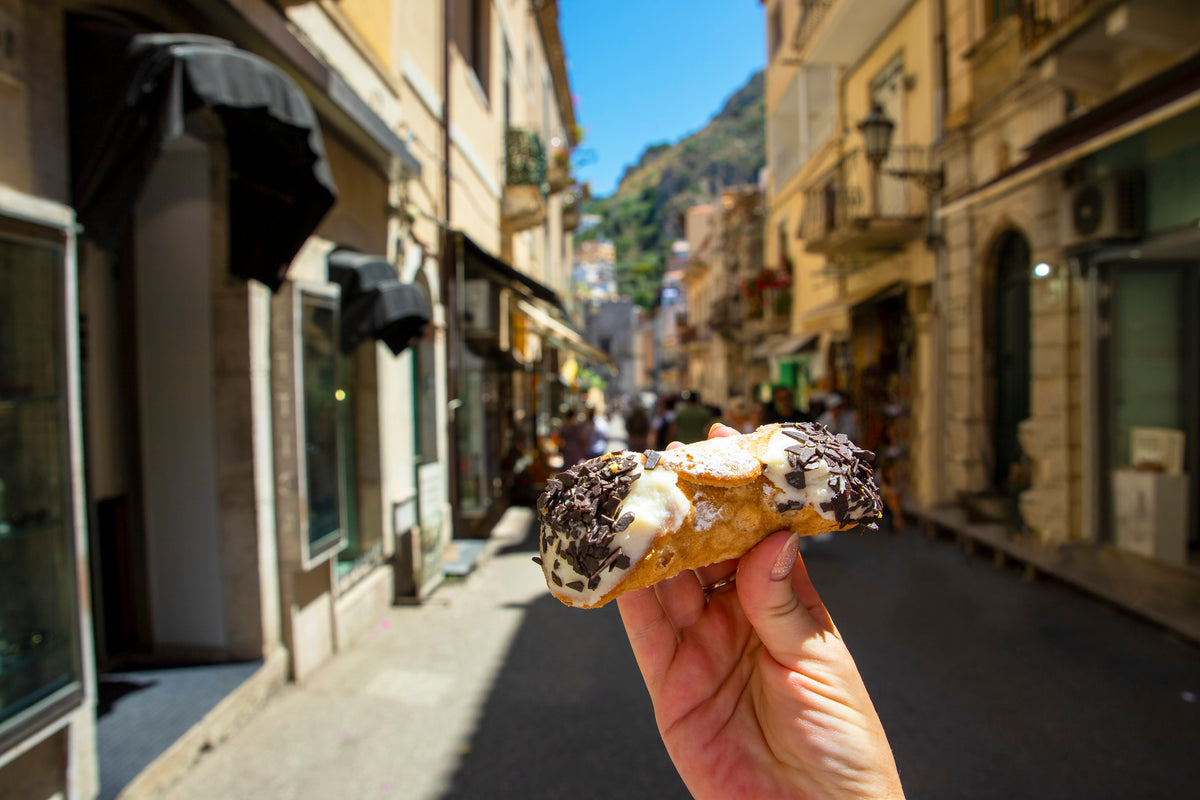 A hand holds a chocolate chip cannoli in the foreground, with a sunny street and old buildings in an Italian town blurred in the background.