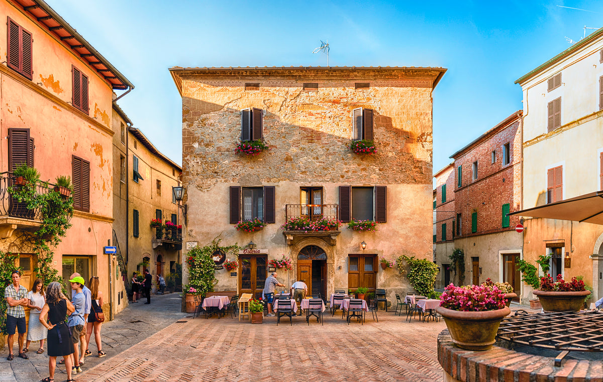 A sunlit Italian piazza features a rustic building with flower boxes, outdoor café tables, and people strolling or chatting. The scene is warm and inviting under a clear blue sky.