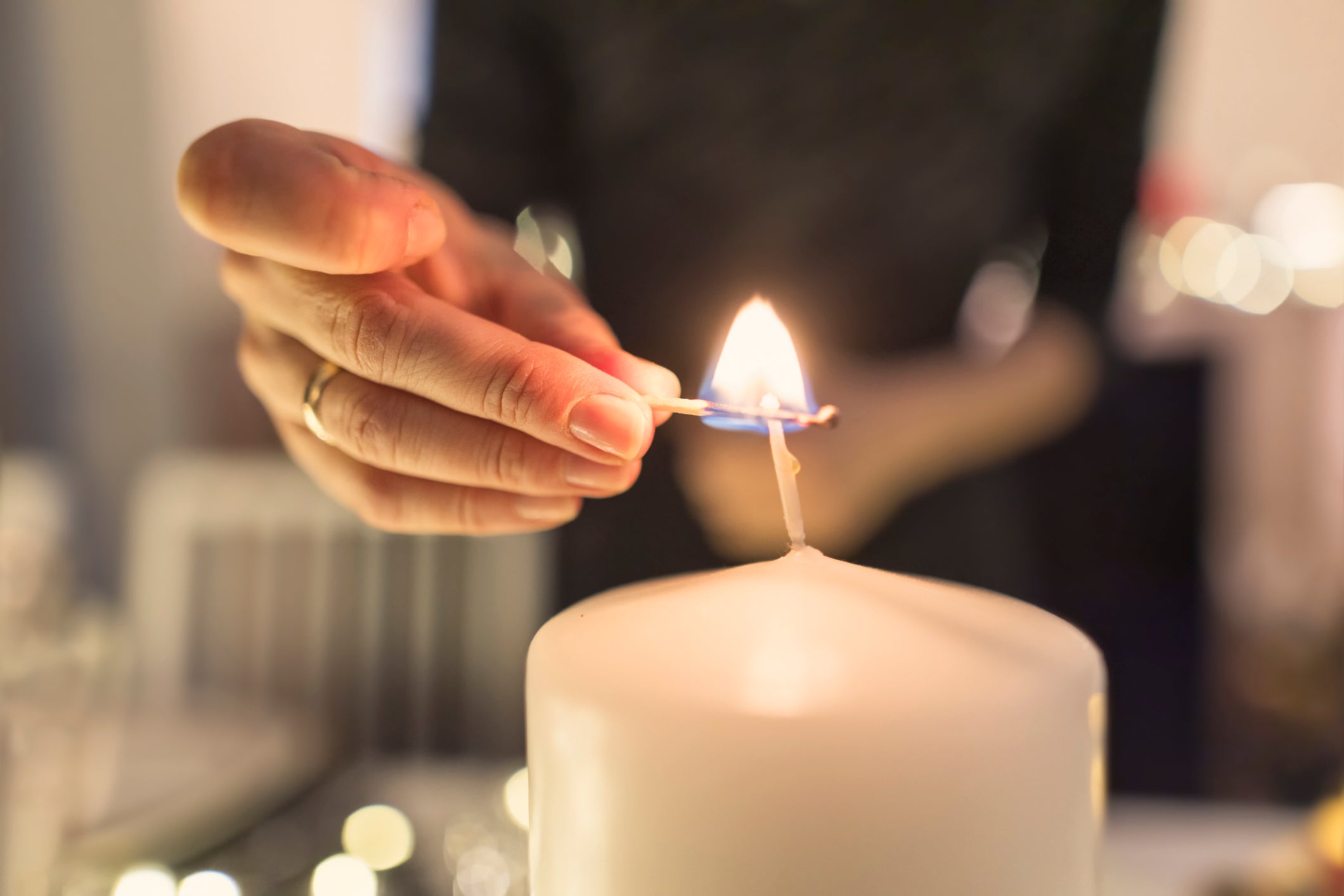 A close-up of a hand lighting a large white candle with a matchstick. The flame is bright, and the background is softly blurred.