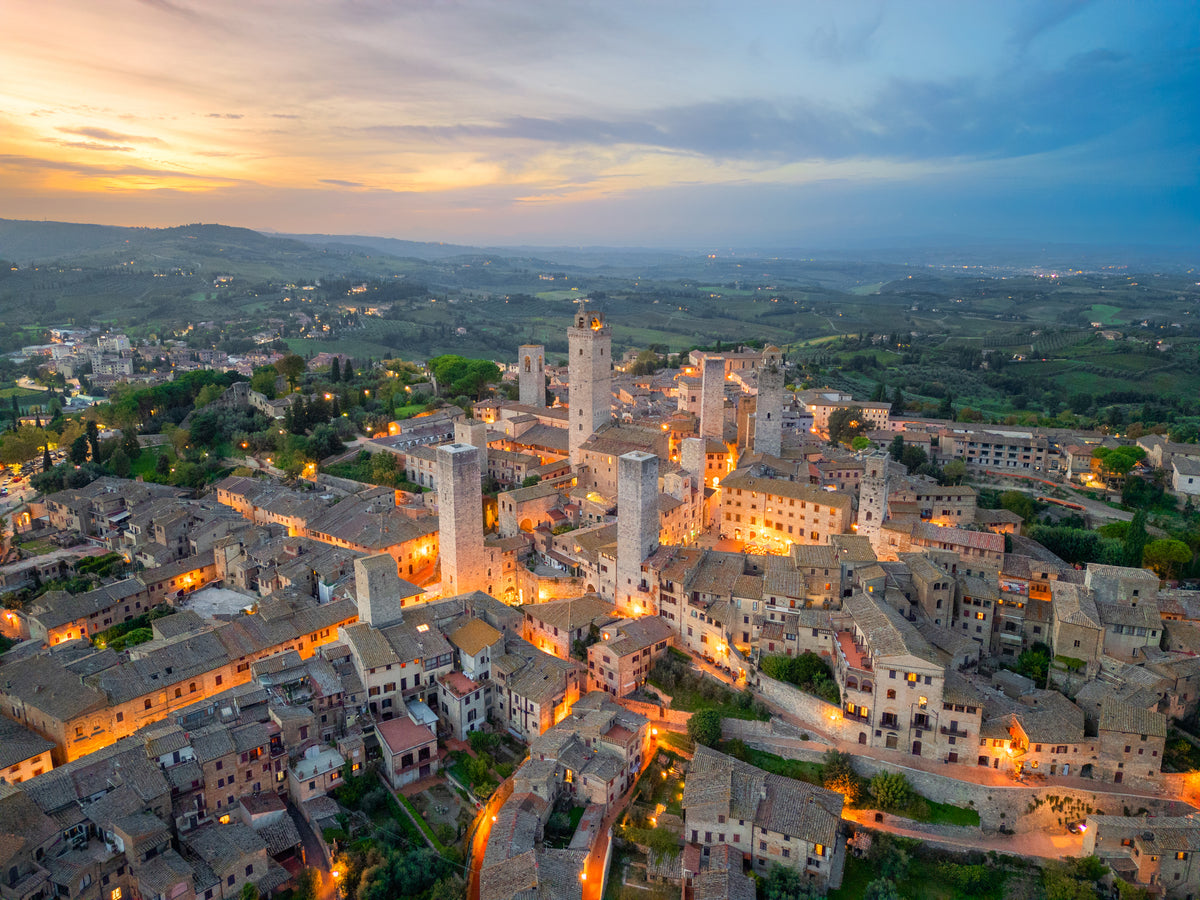 Aerial view of San Gimignano, Italy, at sunset with medieval towers, stone buildings, and surrounding green landscape softly illuminated by streetlights.