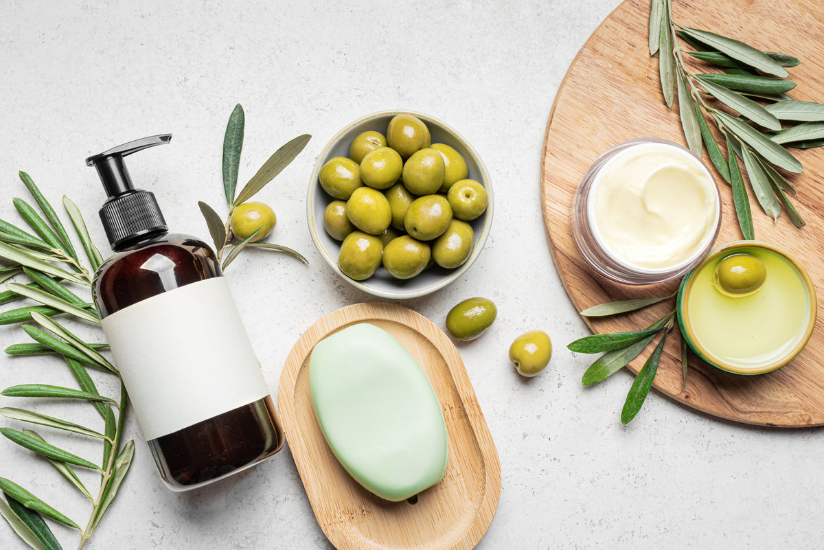 A bottle with a pump, a green bar of soap, a bowl of green olives, two jars of cream, and olive branches are arranged on a light surface with a wooden board.
