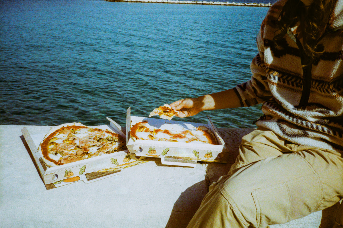 A person sitting by the water, holding a slice of pizza with two open pizza boxes on a concrete surface beside them.